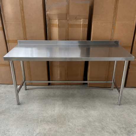 A stainless steel workbench with a rectangular metal top, backrest, and four legs, photographed in a warehouse setting with cardboard boxes in the background.