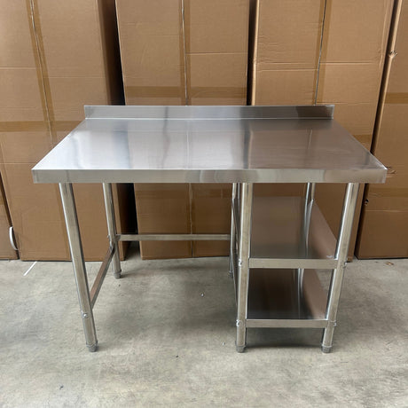 A stainless steel utility table with a rectangular worktop and two open shelves, photographed in a warehouse setting with cardboard boxes stacked on the left and a concrete floor on the right.